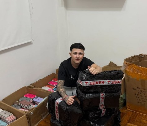 Thiago con pedidos embalados en bolsas negras con cinta de frágil, junto a cajas de fundas